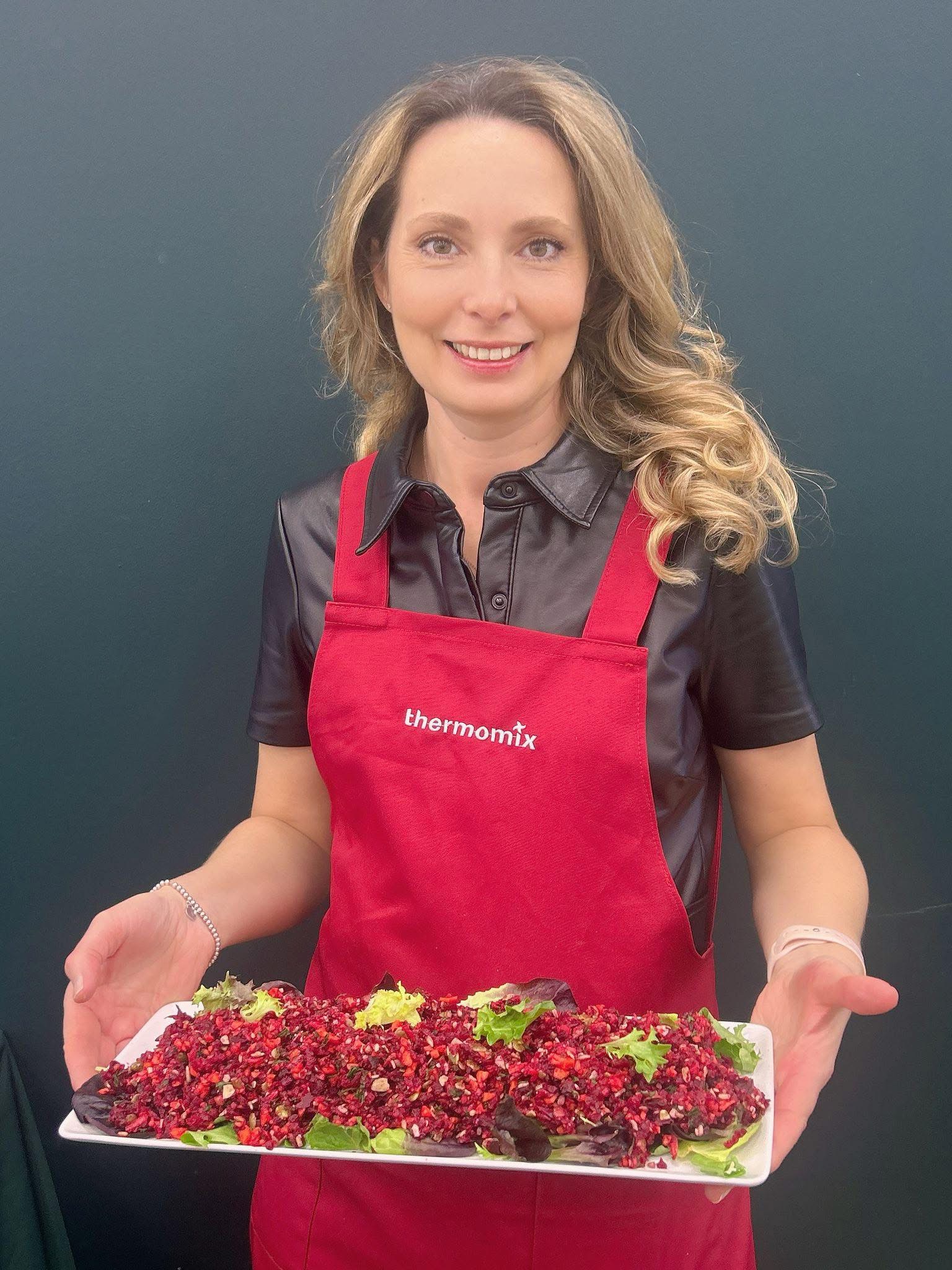 Irina Bratz smiling in her Thermomix apron, holding a fresh beetroot and grain salad